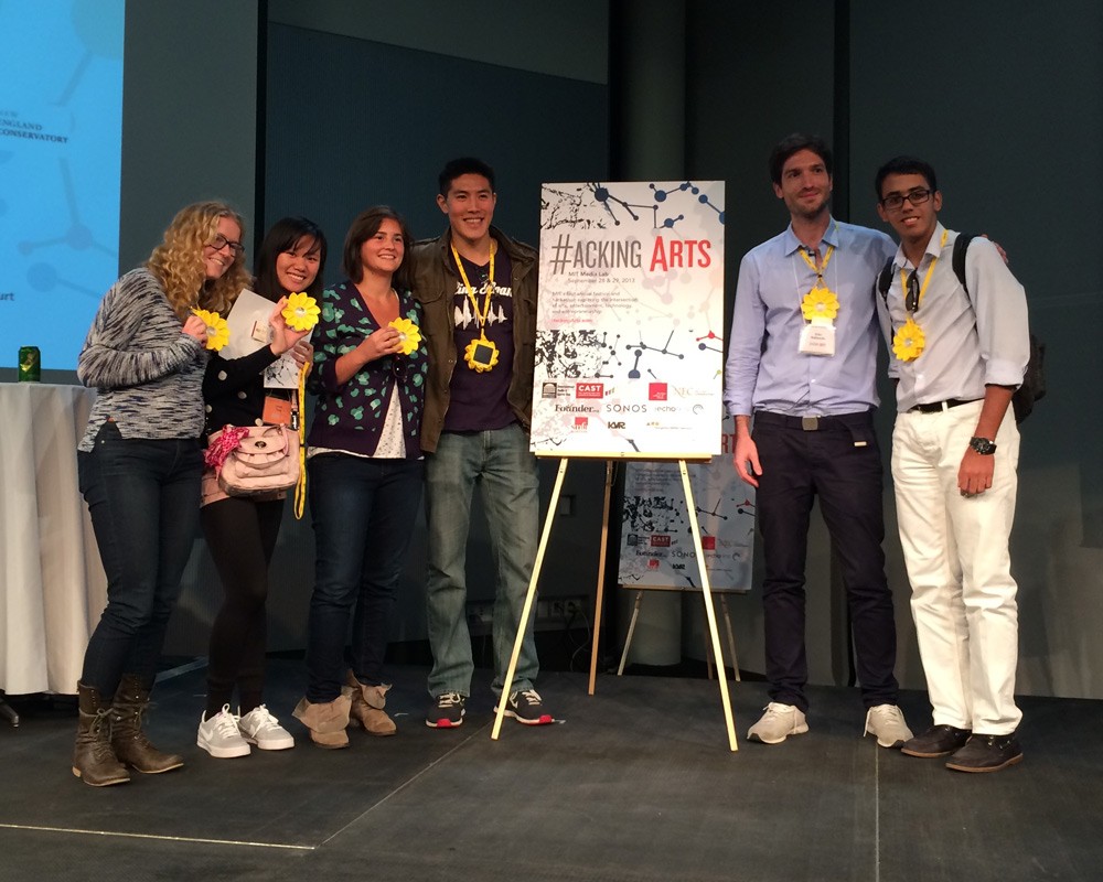 Four people wearing sun-shaped lights on lanyards pose next to a sign reading "Hacking Arts"