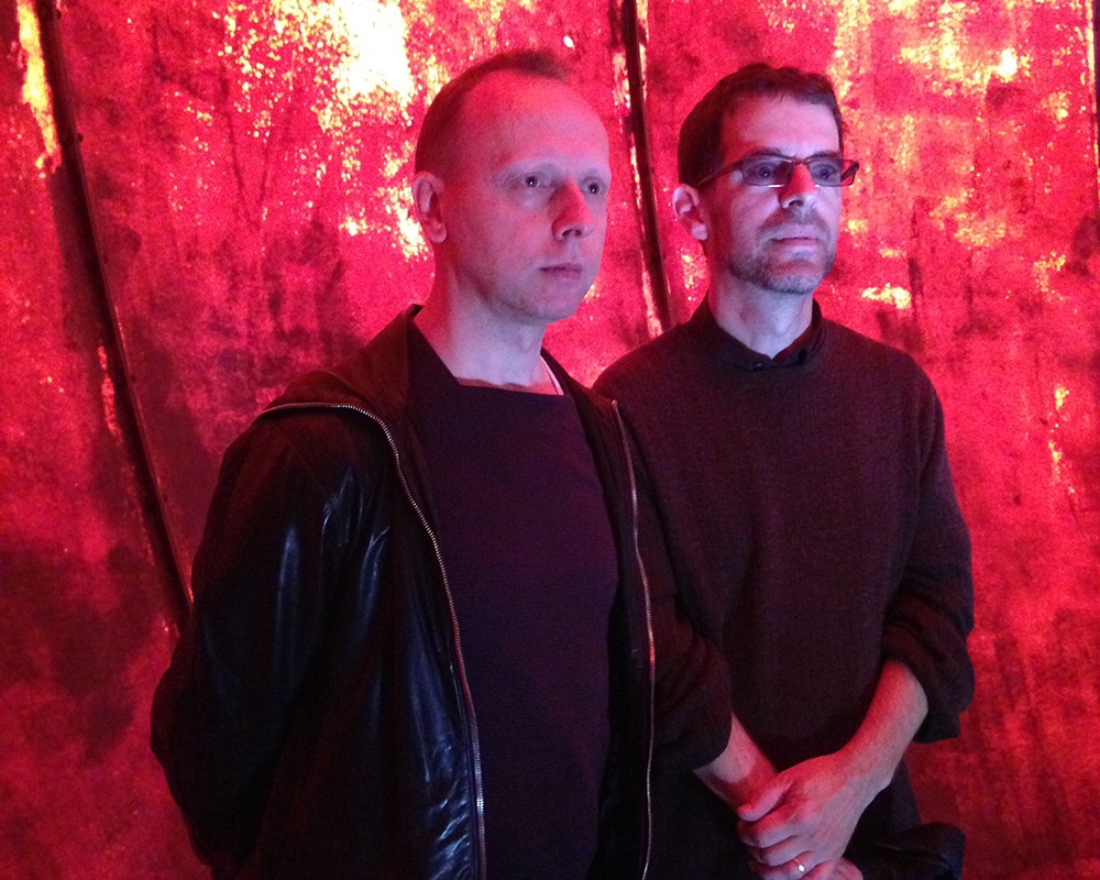 Two men stands against a non-uniform red background