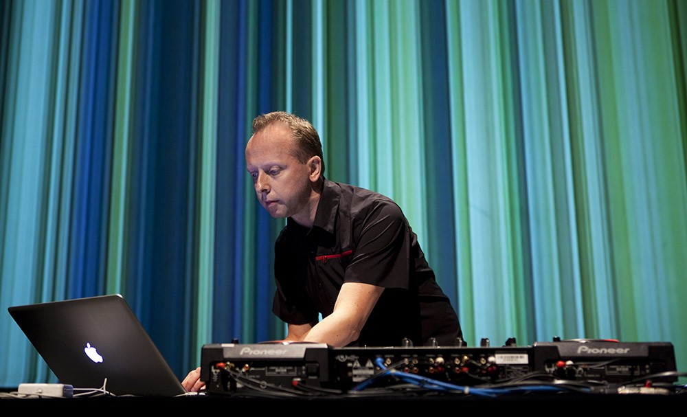 A man works at a laptop and sound board in front of an image of many green and blue vertical lines