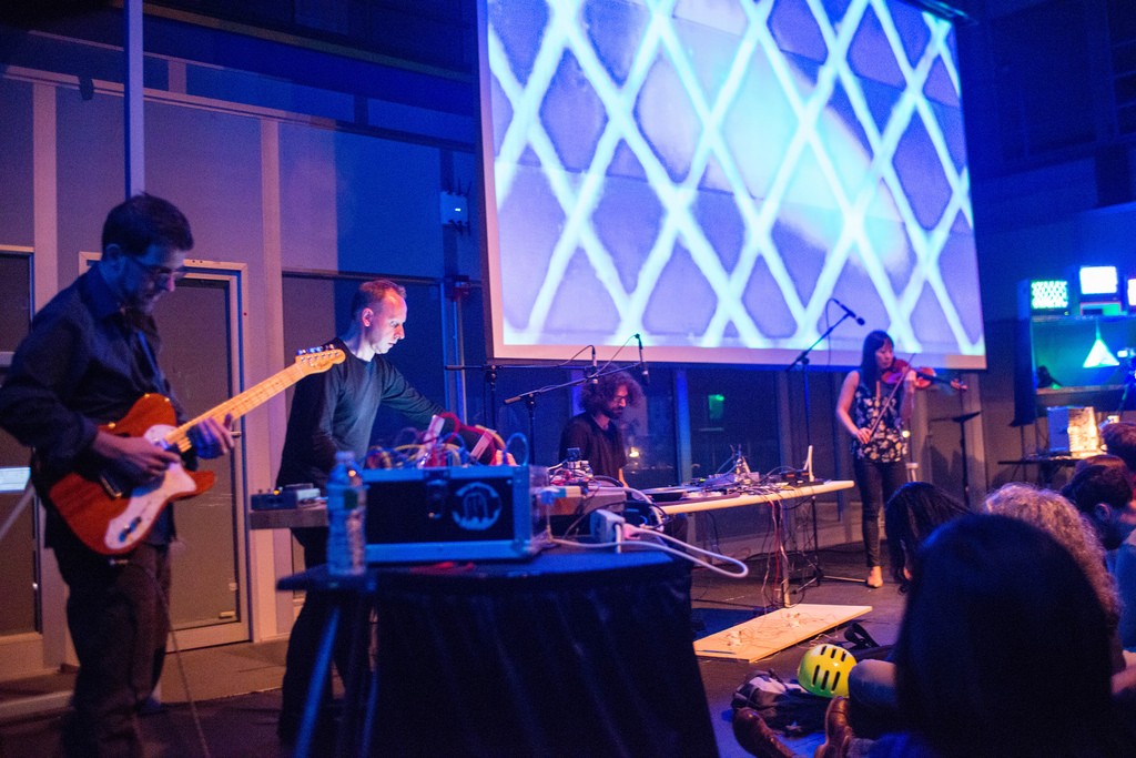 Musicians playing guitar, violin, and electronics perform in front of a projected image.