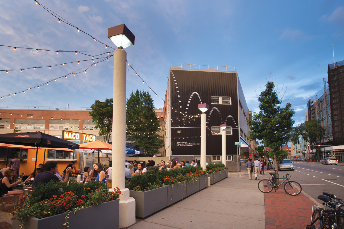 A sidewalk scene with a restaurant, flowers, and decorative lights.