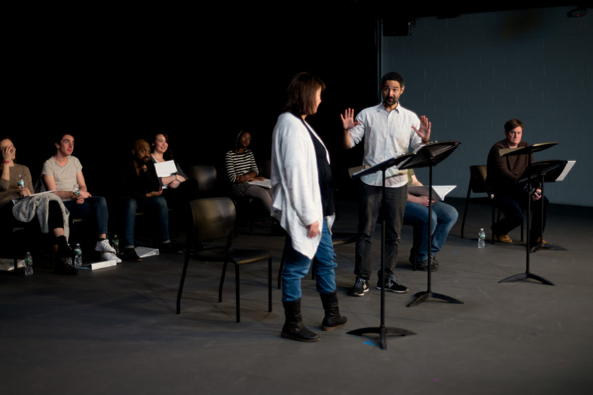 Two actors read from scripts while others look on.