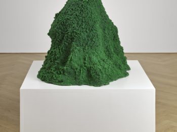 A mountain-like green mound of colored sand, gold, glitter, and crystals sits on a white pedestal in an art gallery.