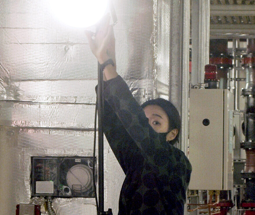 Kwan Queenie Li looks at the camera over her shoulder while adjusting a light fixture aimed at the camera.