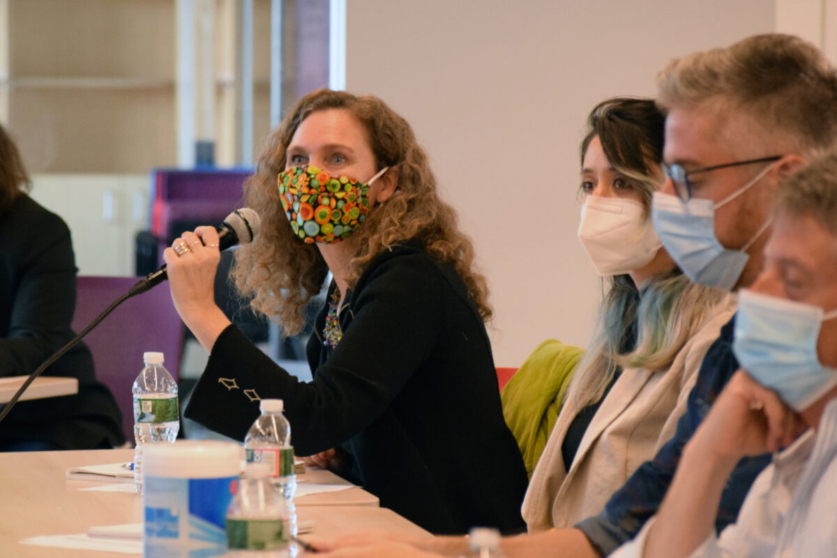 Jen Guillemin speaking with a microphone in a colorful mask and a black blazer.