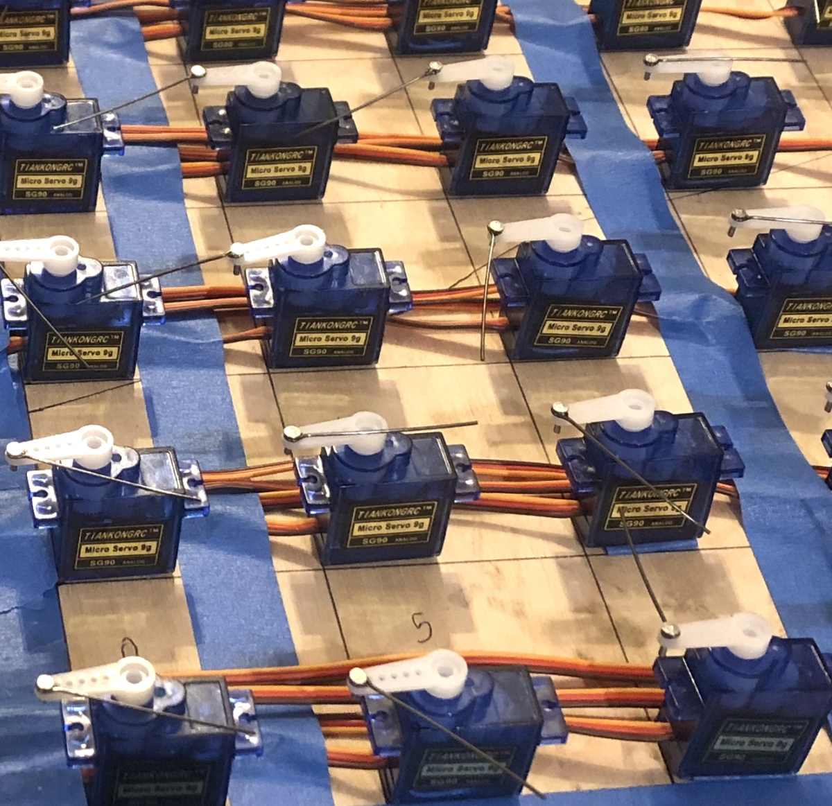 Close up view of more than a dozen electronic parts taped to a wooden board in a grid and connected with red wires.