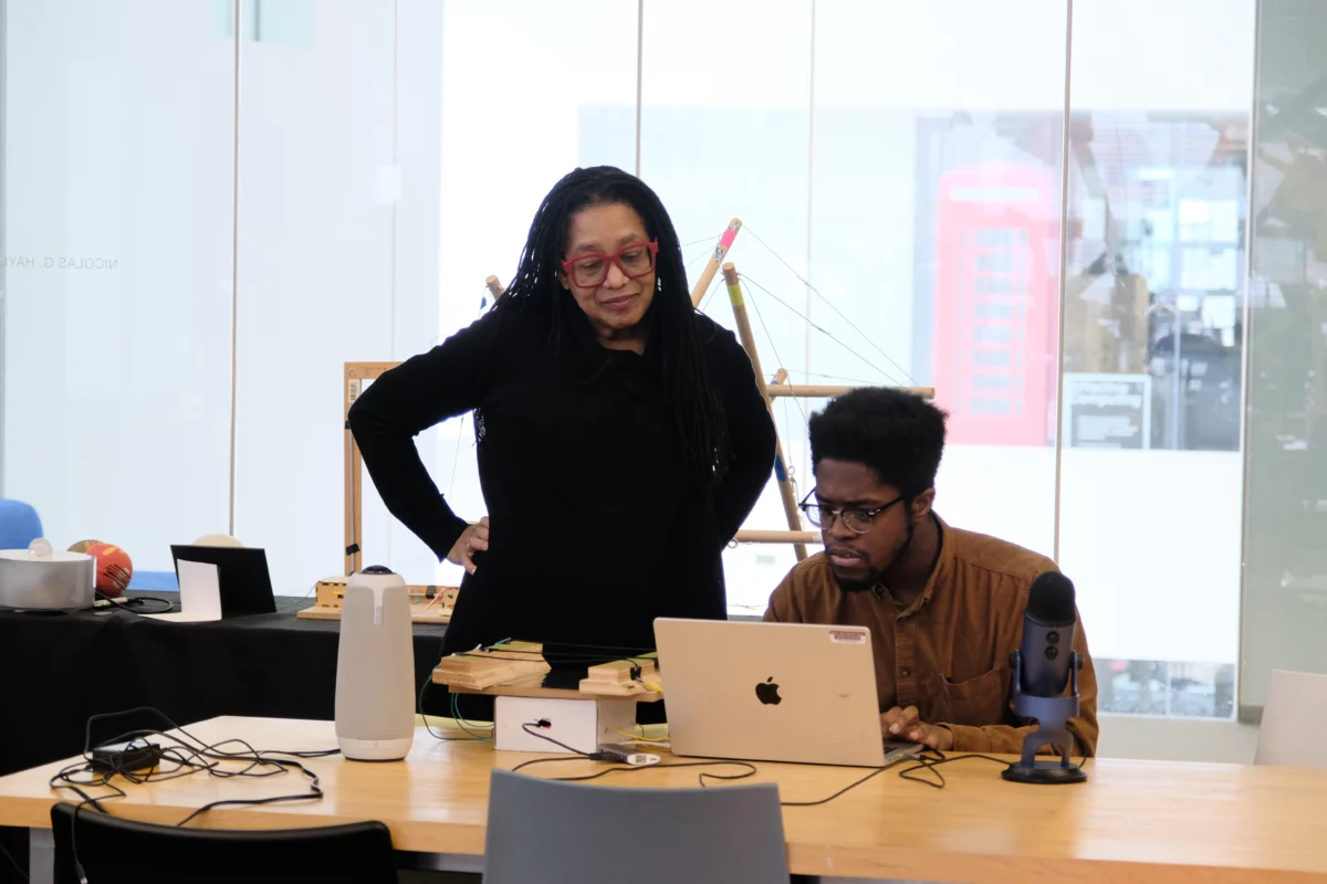 Pamela Z with Max Addae. Addae is seated at a laptop and various electronics, while Pamela Z stands, overlooking and engaging with Addae. Credit Heidi Erickson.