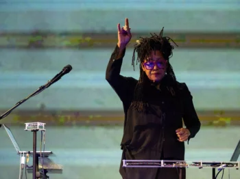 Pamela Z stands with one arm raised, in front of electronics, several laptops, and a microphone wearing black clothing.