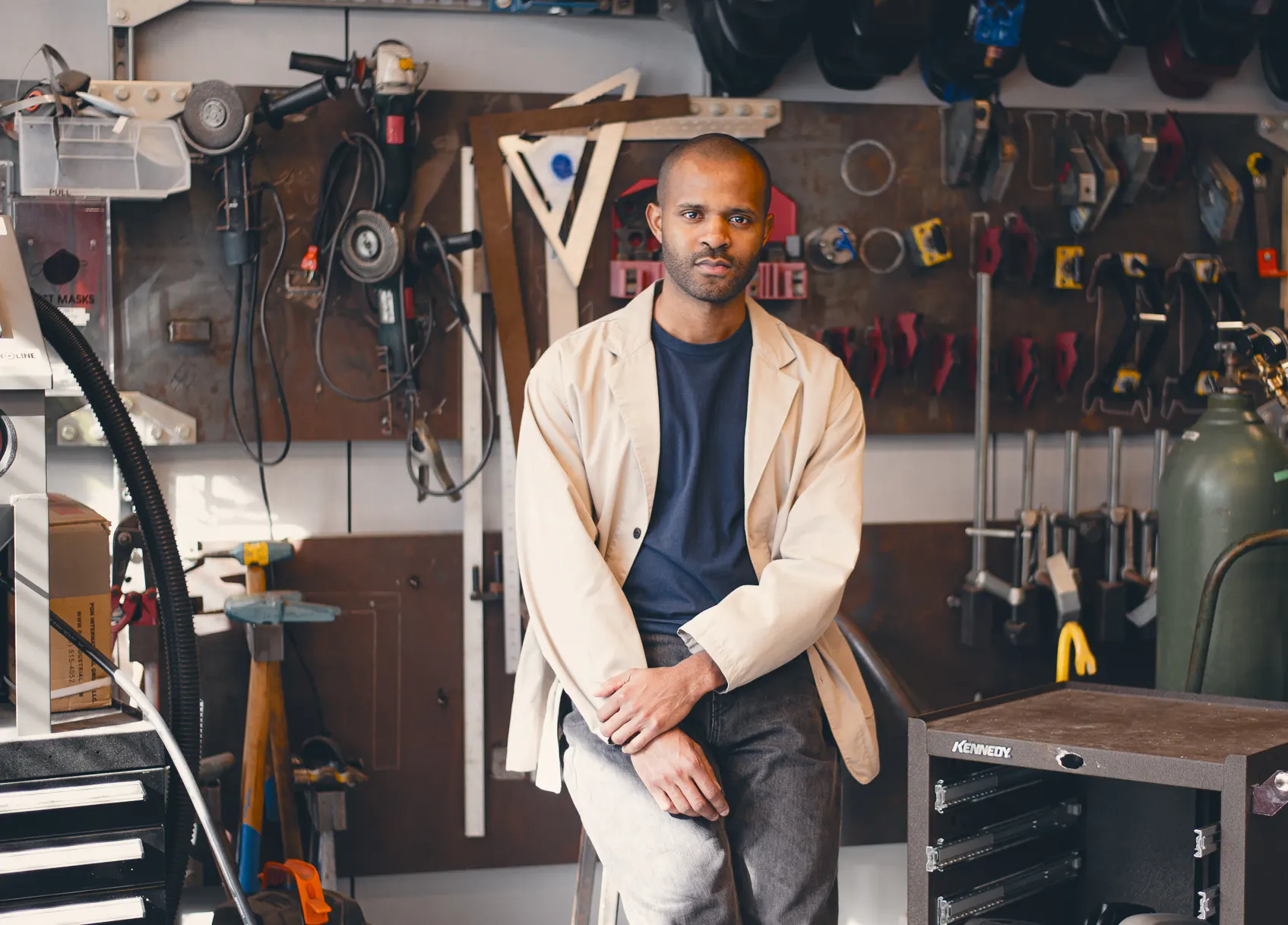 Christopher Joshua Benton looks directly into the camera with arms crossed, while standing in front of an artwork with spokes of lines radiating from the left side of the frame.